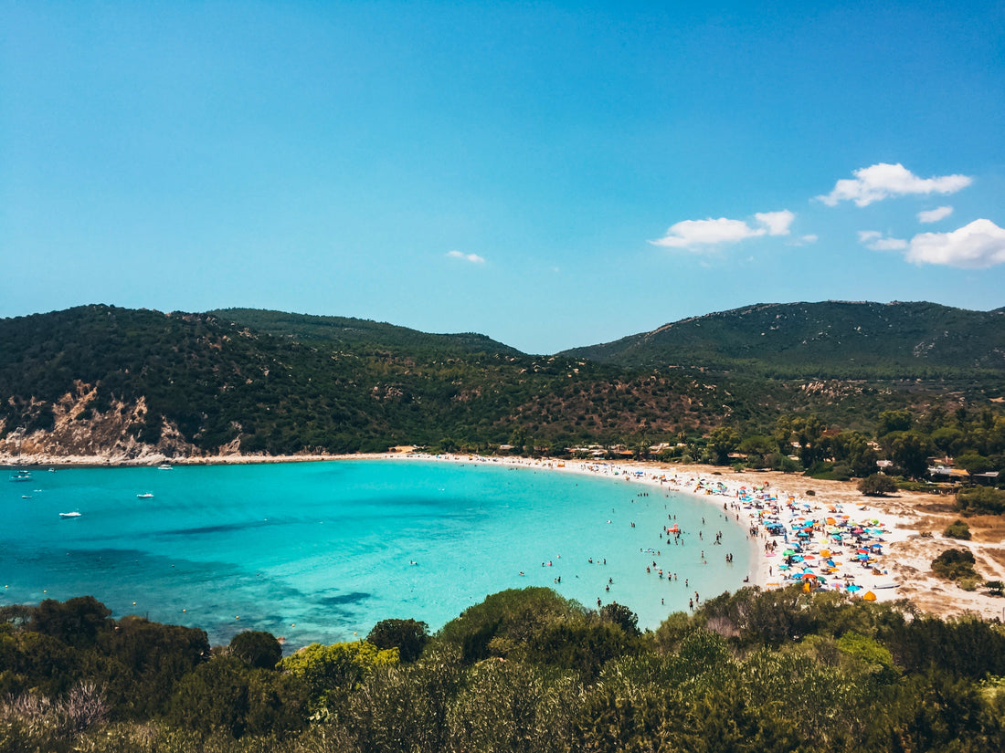 Blue Water Bay With Busy Beach. A busy beach dotted with tourists and a bright blue bay surrounded by low, tree-covered mountains.